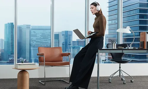 A professional woman wearing white Sony 1000X The Collexion over-ear headphones in an office and city view.