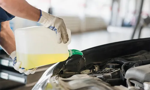 A person pours a bright yellow fluid into a car engine reservoir at an auto shop.
