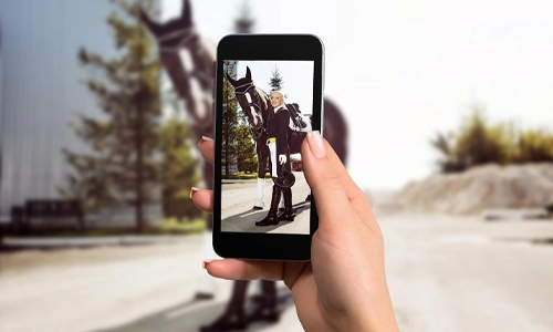 Image of female hands holding mobile phone with photo camera mode on the screen. Picture of woman rider with a horse.