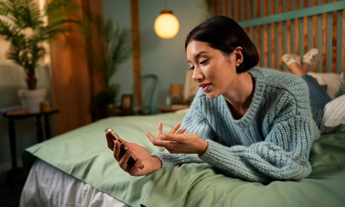 A person sitting in bed holding a smartphone while talking on a video call late at night, illustrating common screen distractions before sleep